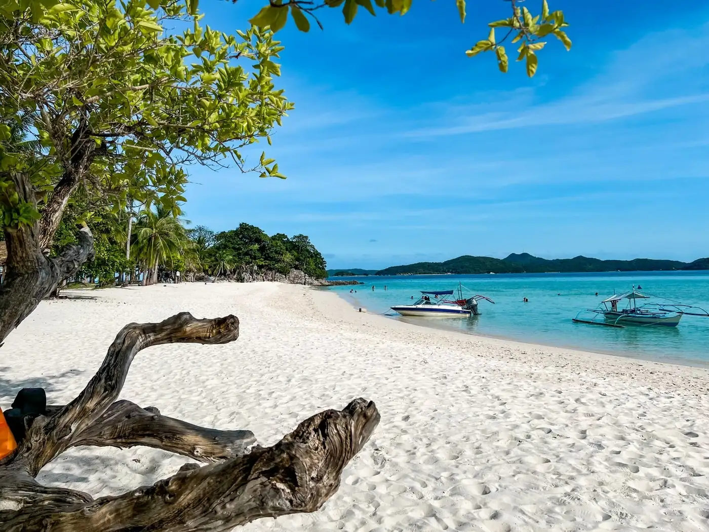 A quiet white-sand beach with turquoise water and boats anchored just offshore, reachable only by water