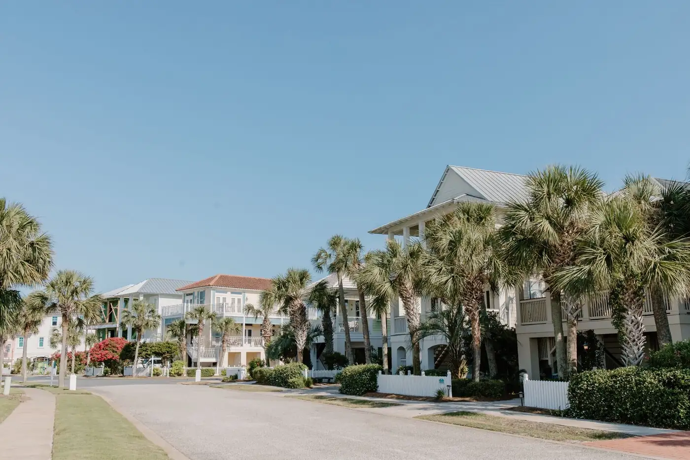 A white clapboard cottage with green shutters behind a picket fence, framed by palms on a quiet Old Naples street